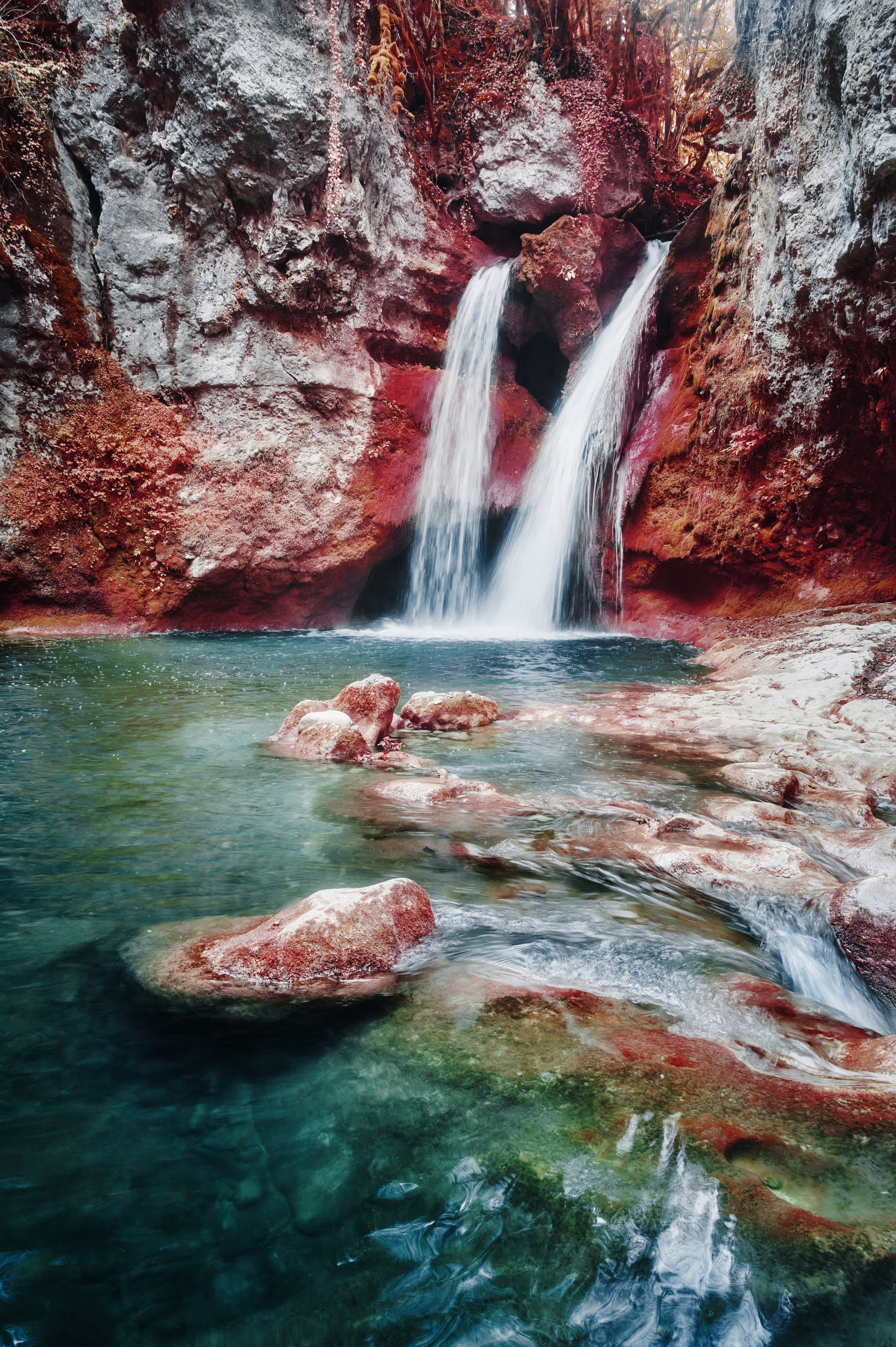 Cascade spectaculaire coulant sur des roches rouges intenses dans un bassin d’eau turquoise. Photographie infrarouge Collection Lumina par Jonas Hangartner, Suisse.