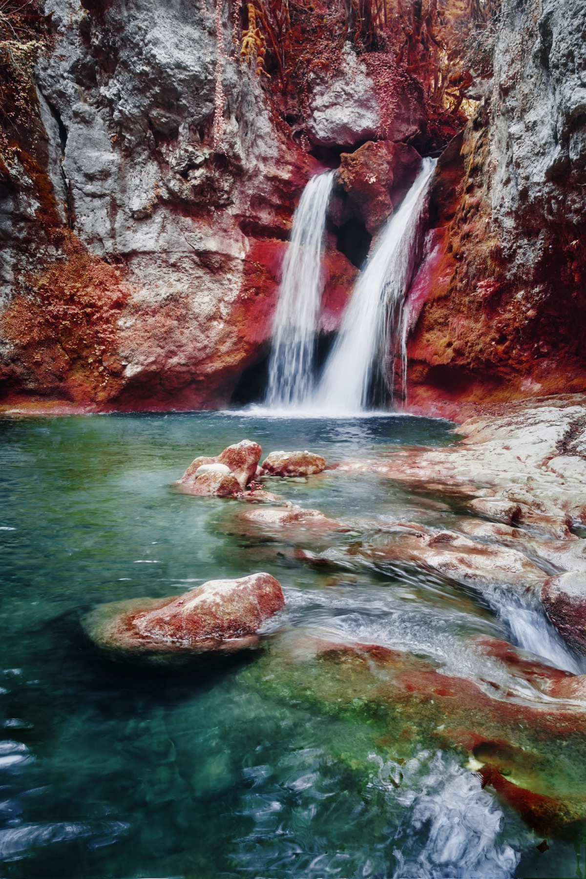Gem : une cascade entre des rochers et une végétation rouge, au-dessus d’un bassin turquoise.